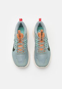Sneaker in tessuto grigio testurizzato con linguetta arancione e dettagli in verde menta. Presenta un logo nero, suole chiare e macchie multicolori. Fettuccia rossa sul tallone.