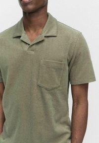 Oliivivärinen lyhythihainen polo-paita pehmeästä kankaasta, jossa on terävä kaulus ja suorakulmainen rintatasku.