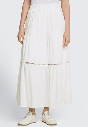 Vrouw die een lange witte rok draagt met kanten inzetstuk en elastische tailleband, gecombineerd met witte lage sneakers op een effen achtergrond.