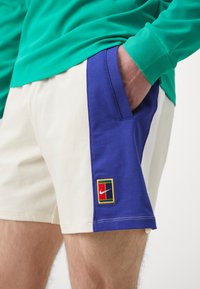 Nike rövidnadrág krém színű anyagból, kék oldalsó panellel és zsebbel, texturált felülettel, valamint vörös, sárga és fekete logóval.