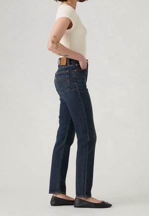 Person in weißem Kurzarm-Oberteil, trägt dunkelblaue Jeans mit geradem Bein und schwarze Ballerinas, steht im Profil mit einer Hand in der Tasche.