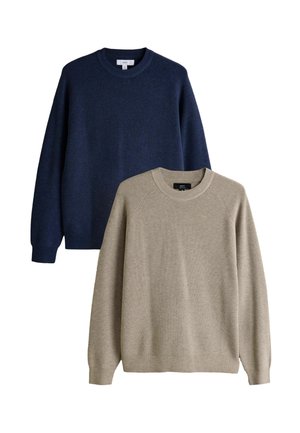 Jonge man staat met handen in de zakken, draagt een beige trui, wit overhemd, marineblauwe broek en witte sneakers tegen een effen achtergrond.