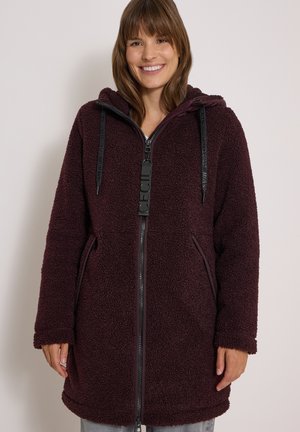 Femme portant une longue veste en polaire violet foncé avec une fermeture éclair et une capuche, souriant sur un fond clair uni.