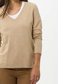 Pull beige à col en V en matière tricotée douce avec manches longues et poignets côtelés, superposé sur une chemise blanche. Détail subtil de texture.