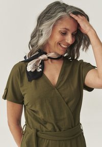 Abito avvolgente verde oliva con maniche corte e vita annodata. Abbinato a una sciarpa in seta nera e crema con un motivo floreale.