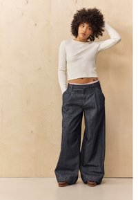 Pantalones de mezclilla de pierna ancha en azul oscuro con un acabado texturizado. Combinados con una camiseta de manga larga de canalé ajustada en color crema. Estilo relajado y casual.