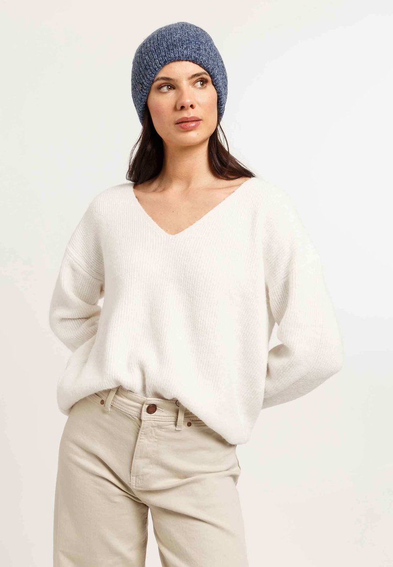 Fehér kötött pulóver V-kivágással, világos bézs nadrággal párosítva. Egy kék kötött sapka teszi teljessé az öltözéket. Sima textúra, laza szabás.