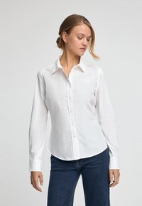 White long-sleeve button-up shirt made of smooth cotton. Features a classic collar, rounded hem, and subtle logo on the chest.