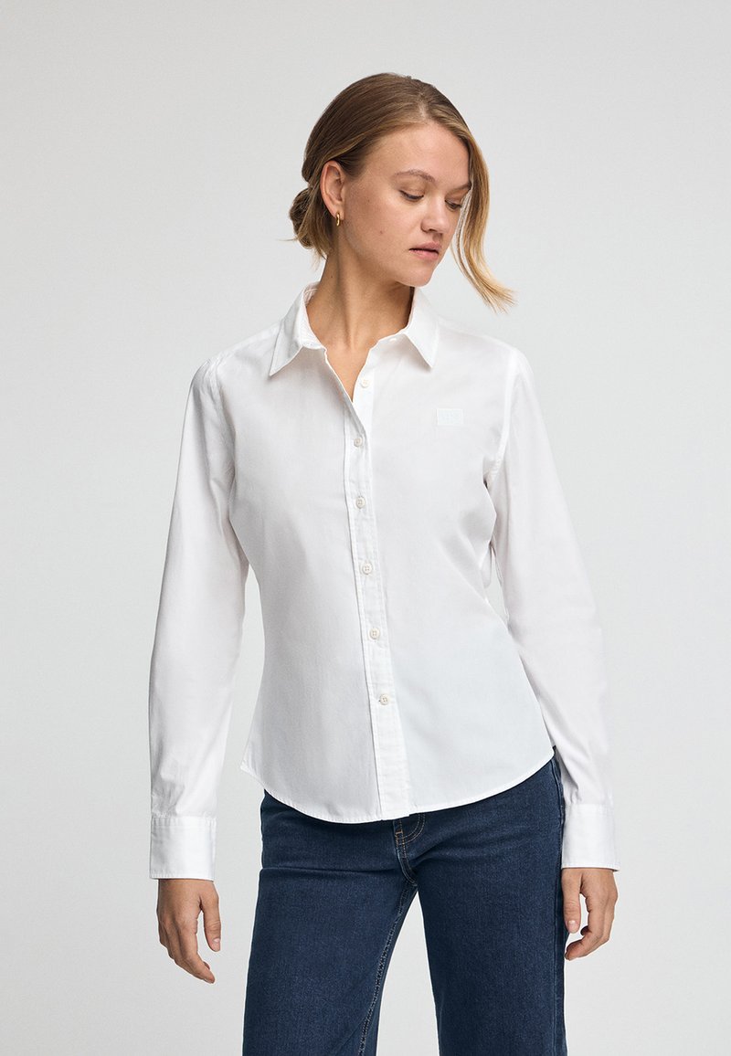White long-sleeve button-up shirt made of smooth cotton. Features a classic collar, rounded hem, and subtle logo on the chest.