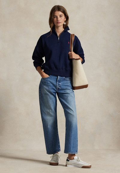 Mujer que lleva una sudadera azul marino con cremallera de cuarto de largo, jeans azul claro, zapatillas blancas, y lleva un bolso de tela beige con correas marrones, de pie frente a un fondo neutro.