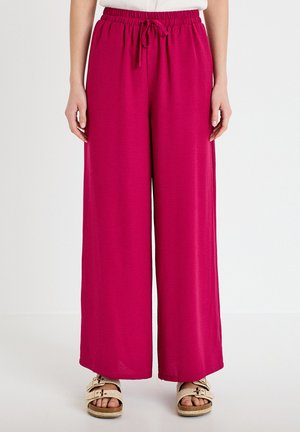Donna che indossa pantaloni a gamba larga a vita alta di colore rosa acceso con coulisse in vita e sandali beige aperti sulle dita, su sfondo bianco.