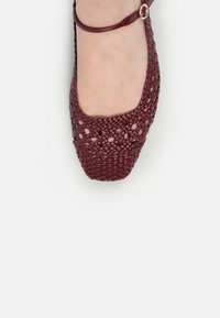 Sandală plată din piele burgundă împletită, cu curea la gleznă, purtată pe un picior, evidențiind un detaliu de model împletit sub formă de plasă și vârf pătrat.