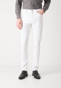 Bijele slim-fit traperice s pet džepova, srebrnim detaljima i kontrastnim šavovima, uparen sivoj košulji na dugmad i crnim loafers cipelama.