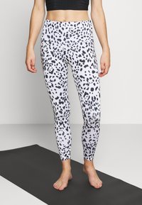White leggings with a black spotted pattern, featuring a high waistband. The model stands barefoot on a black yoga mat.