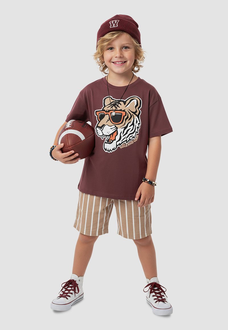 Smilende lille barn iført en mørkerød hat, t-shirt med tigerprint, stribede shorts og sneakers, holder en amerikansk fodbold ved siden.