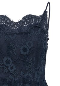 Gros plan d’un caraco en dentelle noire avec des motifs floraux, des bords festonnés et de fines bretelles réglables sur fond blanc.