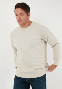 Sudadera de color beige claro con mangas largas raglán y cuello redondo. Presenta un pequeño logo en el pecho y un tejido suave y texturizado.