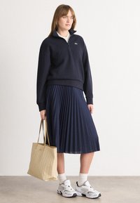 Navy zip-up sweatshirt, pleated navy skirt, white socks, sporty white sneakers with black accents, and a textured beige tote bag.
