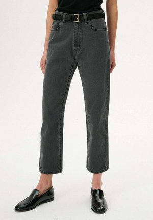 Osoba ubrana w czarne, wysokie spodnie dżinsowe z podciętymi nogawkami, czarny pasek i czarne skórzane buty wsuwane, stojąca na jednolitym tle.