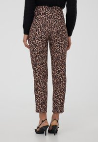 Donna che indossa pantaloni a stampa leopardata a vita alta e aderenti, con tacchi alti neri aperti sul retro, in piedi contro uno sfondo neutro.