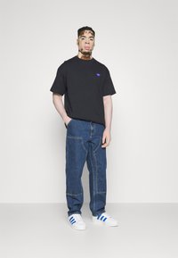 Schwarzes Baumwoll-T-Shirt mit kurzen Ärmeln und kleinem lila Logo auf der Brust, kombiniert mit lockeren blauen Jeans und weißen Sneakers mit blauen Streifen.