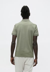 Mężczyzna z dredami, ubrany w zieloną koszulkę polo i jasnoszare spodnie, stojący tyłem na jednolitym białym tle.