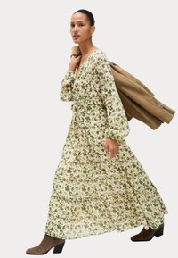 Langes, blumiges Kleid mit grünen und weißen Blumen, gestuftem Design, taillierter Passform und langen Ärmeln. Brauner Mantel über der Schulter getragen. Stiefel mit Absatz.