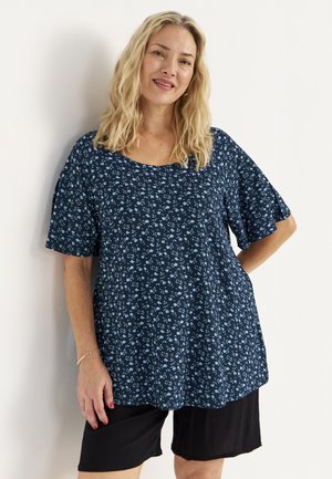 Blonde Frau lächelt, trägt ein blaues, geblümtes Kurzarmshirt und schwarze Shorts bis zum Knie, steht vor einem weißen Hintergrund.