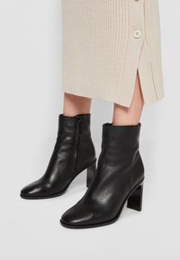 Stivali neri in pelle fino alla caviglia con punta affusolata e tacco a blocco spesso. Chiusura con cerniera laterale e texture liscia, abbinati a un vestito in maglia beige.