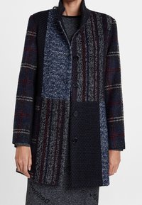 Abrigo de mezcla de lana con diseño de patchwork en azul oscuro, gris y burdeos, con texturas distintivas, cuello y cuatro botones negros.