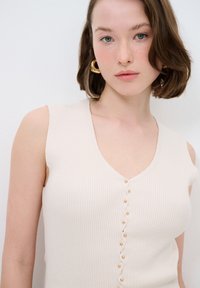 Hellbeige, geripptes Tanktop mit V-Ausschnitt, das vertikale Streifen und goldfarbene Knöpfe an der Vorderseite aufweist. Glatte, enganliegende Textur.