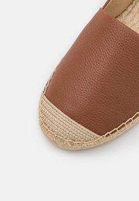 Chaussure espadrille en cuir marron avec bout en jute tissé et bord de semelle en jute tressée sur fond blanc.