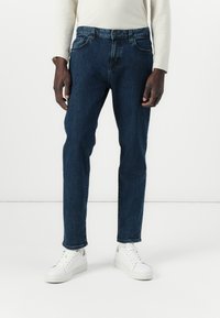 Jeans de mezclilla azul oscuro de corte ajustado con cinco bolsillos, que presentan un cierre de cremallera y botón, combinados con zapatillas blancas.