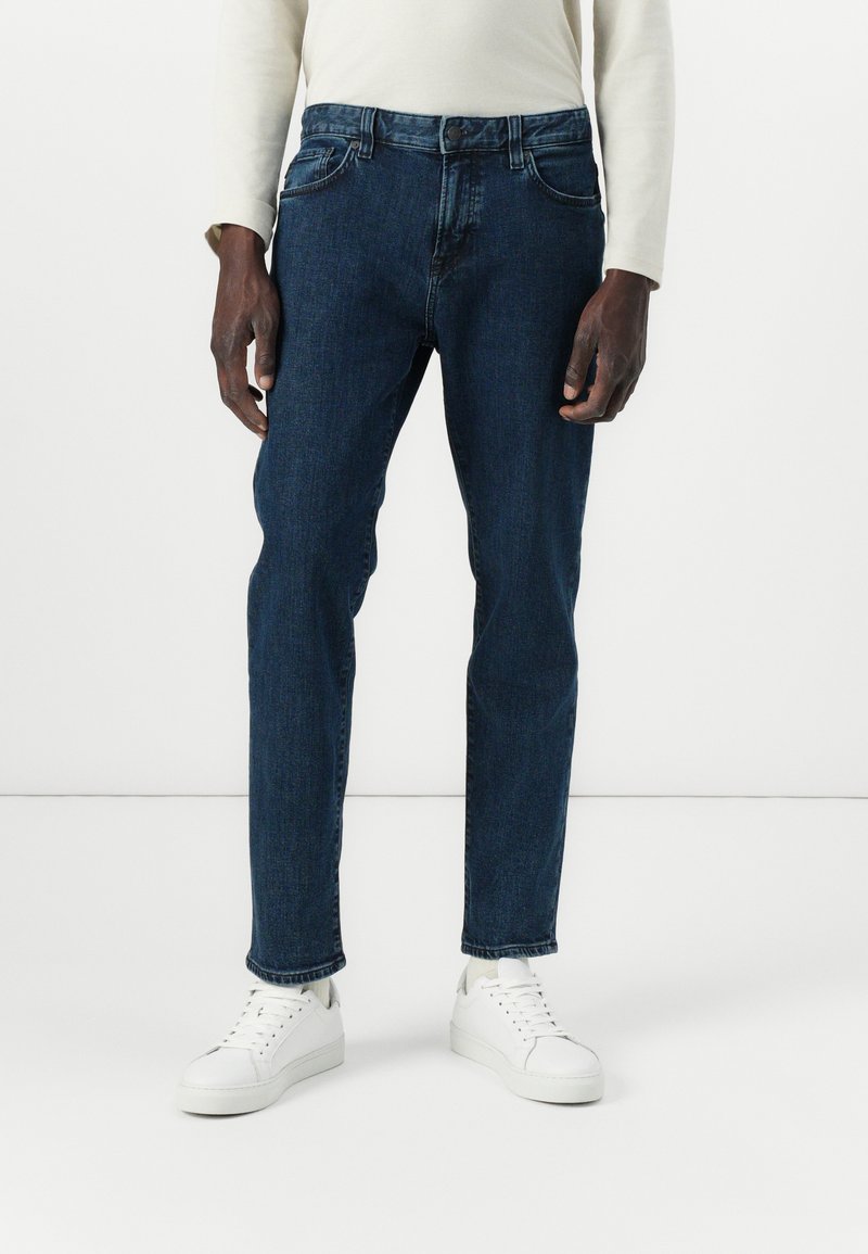 Jeans de mezclilla azul oscuro de corte ajustado con cinco bolsillos, que presentan un cierre de cremallera y botón, combinados con zapatillas blancas.
