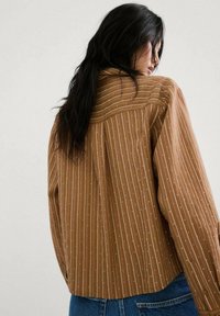 Mujer de cabello largo y oscuro, vestida con una camisa marrón a rayas finas con pequeños puntos blancos, vista desde atrás sobre un fondo liso.