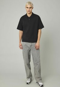 Polo noir avec un col et des manches courtes, associé à un jeans gris clair. Le modèle porte des baskets blanches avec des accents noirs.