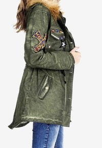 Parka vert olive avec col en fourrure, patches décoratifs, accents métalliques et poches zippées. Manches longues et forme légèrement ajustée.