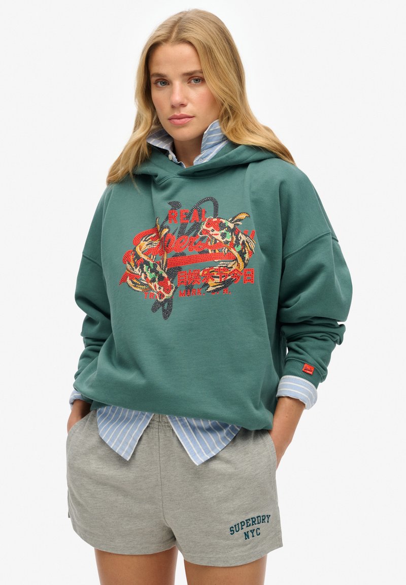 Sweatshirt à capuche vert avec un design de dragon brodé rouge et multicolore, accompagné d'un short gris affichant le texte "SUPERDRY NYC". T-shirt rayé en dessous.