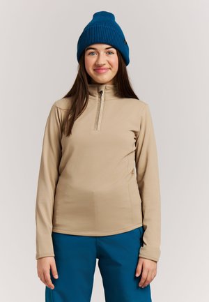 Junge Frau, die einen beigen Reißverschluss-Pullover, blaue Hose und eine blaue Strickmütze trägt, steht vor einem schlichten hellen Hintergrund.