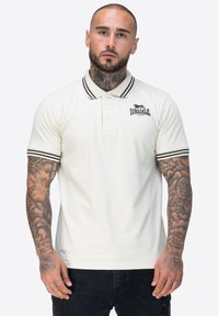 Mann mit Tattoos am Hals und an den Armen trägt ein weißes Lonsdale-Polohemd mit schwarzen Streifen am Kragen und an den Ärmeln, steht vor einem einfarbigen Hintergrund.