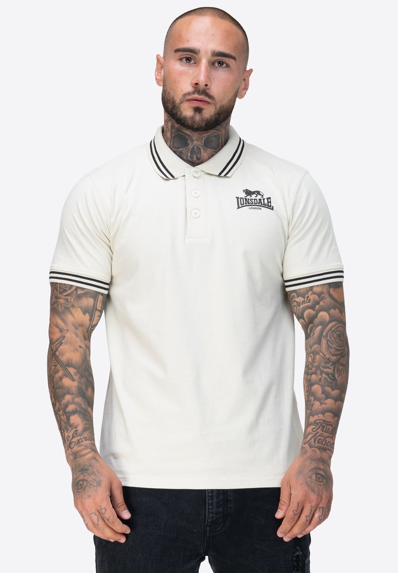 Mann mit Tattoos am Hals und an den Armen trägt ein weißes Lonsdale-Polohemd mit schwarzen Streifen am Kragen und an den Ärmeln, steht vor einem einfarbigen Hintergrund.