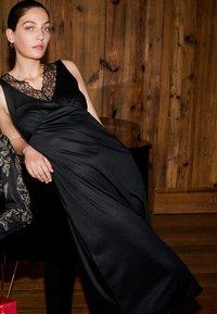 Abito da sera nero con scollatura in pizzo, tessuto fluido e vita aderente. Modella seduta contro uno sfondo di legno, in mostra linee eleganti.