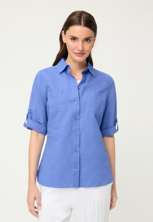 Donna con camicia blu a bottoni con le maniche arrotolate e pantaloni bianchi, in piedi davanti a uno sfondo chiaro uniforme.