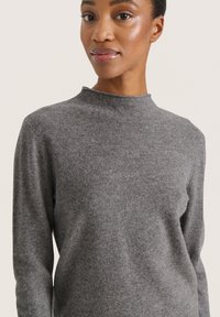 Grauer, langärmeliger Rollkragenpullover aus weichem Material, mit taillierter Passform und subtiler Textur. Nahaufnahme des Ausschnitts.