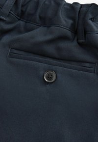 Mörkblått tyg med en slät yta, med en ficka i lapptäcksstil och en svart knapp med "Clarks" ingraverat, placerad vid fickans kant.