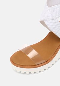 Sandales à plateforme blanches avec un design texturé, dotées d'une sangle en plastique transparent et d'une semelle intérieure beige. Semelle antidérapante avec accents en relief.