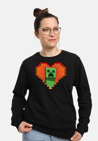Felpa nera con un design in pixel art di un creeper verde a forma di cuore, circondata da dettagli arancioni. Vestibilità casual con scollatura tonda.