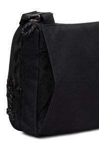 Gros plan sur un coin d'un sac messager en tissu noir avec une bandoulière, une boucle et des détails de couture texturée.