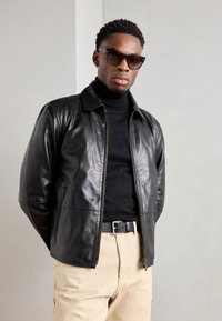 Μαύρο δερμάτινο μπουφάν με δομημένο γιακά και φερμουάρ, φορεμένο πάνω από ένα μαύρο ζιβάγκο, σε συνδυασμό με ανοιχτόχρωμο παντελόνι.