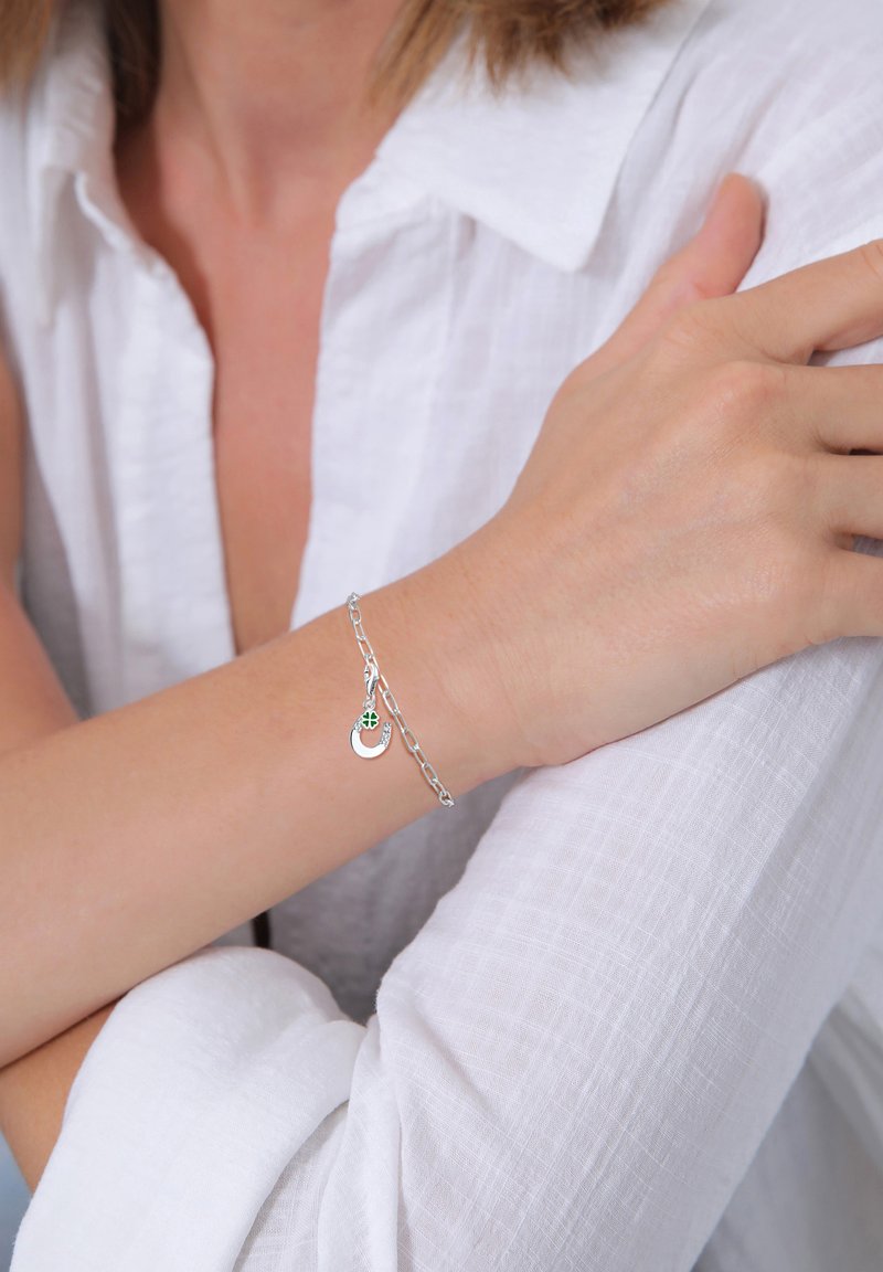 Silberne Kettenarmband mit einem runden Anhänger in grünem Akzent, am Handgelenk getragen, kombiniert mit einem hellen, strukturierten weißen Hemd.